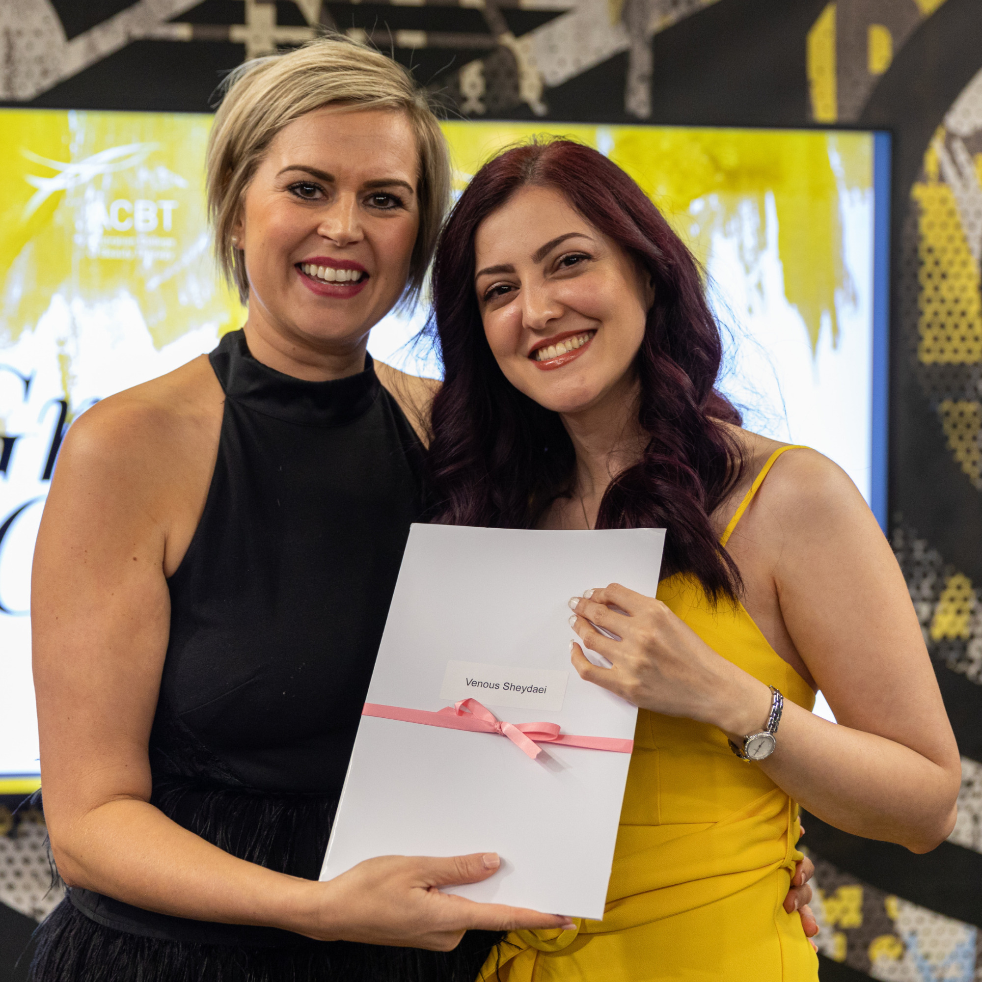 ACBT Graduate, Venus, accepting her certificate from lecturer, Kirsty. Venus is wearing a yellow dress and Kirsty is wearing a black dress. Both ladies are smiling.